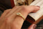 A close-up of a hand wearing a yellow gold ring with a marquise-cut diamond. The ring features a textured band and a diamond-set crown, shown against a background of a wooden box and a cream-colored book.